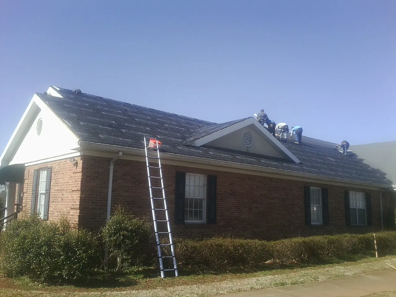 Crew tearing shingles off a brick ranch roof with a ladder set — Emergency Roof Repair work by our crew in Gully